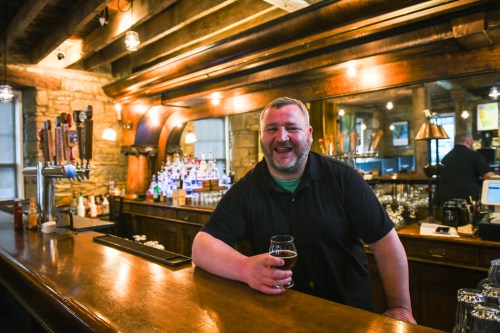 A smiling bartender stands at a wooden bar, holding a glass of beer, with shelves of drinks in the background.