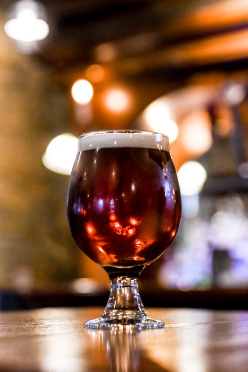 A glass of amber beer with a frothy head, set on a wooden table in a dimly lit bar.