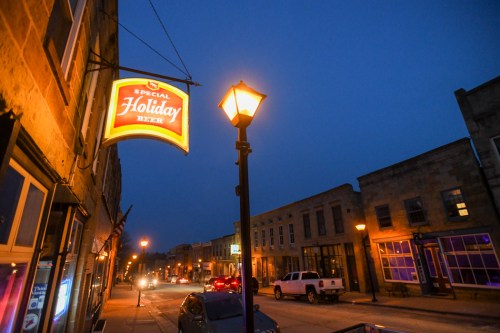 A street scene at dusk featuring a glowing "Special Holiday Beer" sign and lampposts illuminating the quiet road.