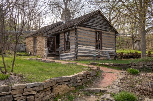 A rustic log cabin with a stone foundation, surrounded by greenery and a stone pathway.