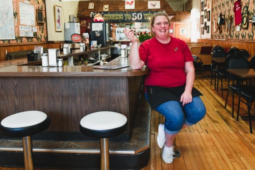 A smiling woman in a red shirt sits at a diner counter, holding a spoon, with a cozy restaurant atmosphere around her.