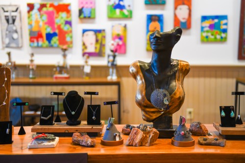 A display of jewelry on a wooden table, featuring a bust and various artistic pieces in the background.