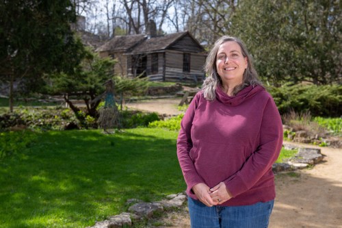 A smiling woman in a maroon hoodie stands in a garden with a rustic cabin in the background.