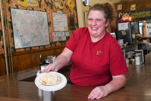 A smiling woman in a red shirt holds a plate of food at a diner, with a colorful wall in the background.
