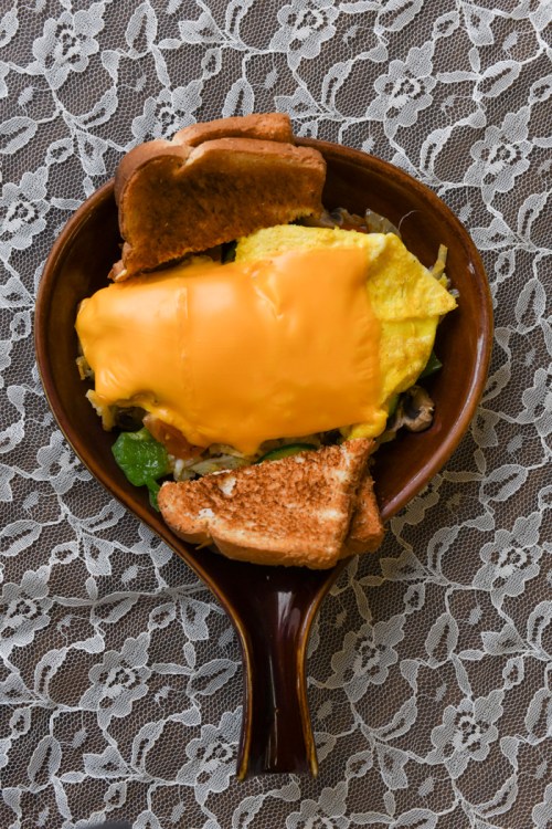 A bowl of omelette topped with melted cheese, surrounded by slices of toast on a lace tablecloth.