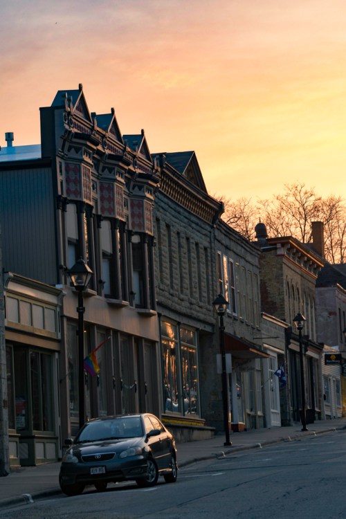 A quiet street lined with historic buildings at sunset, featuring a rainbow flag and a parked car.