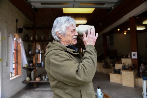 A man with gray hair holds a ceramic mug to his face, smiling in a pottery studio.