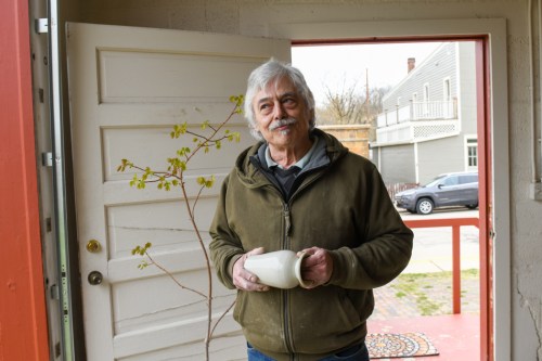 A man with gray hair and a mustache stands in a doorway, holding a small white pot, with a branch nearby.