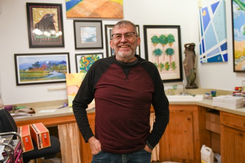 A smiling older man stands in an art studio surrounded by colorful paintings and wooden decor.