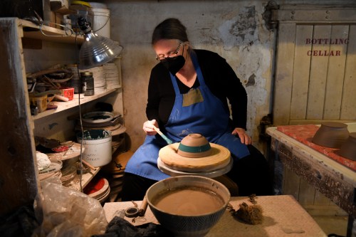 A woman in a blue apron shapes clay on a pottery wheel in a rustic workshop, wearing a black mask.