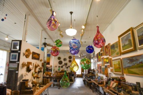 Colorful glass ornaments hang from the ceiling in an art-filled shop, showcasing various paintings and decor.
