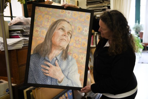 A woman holds a framed portrait of a person with long hair, gazing thoughtfully, surrounded by stacks of papers.