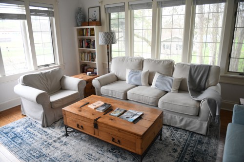 Cozy living room with a light sofa, armchair, wooden coffee table, and large windows letting in natural light.