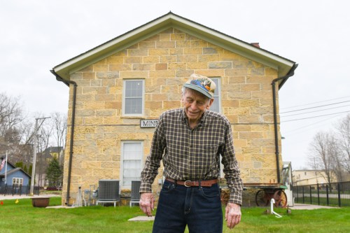 An elderly man in a plaid shirt and cap stands in front of a stone building, smiling slightly on a cloudy day.