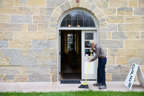 An elderly man sweeps the entrance of a stone building with an "OPEN" sign nearby.