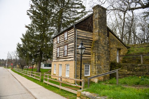 A rustic log cabin with a stone chimney, surrounded by trees, near a quiet road and a lamppost.