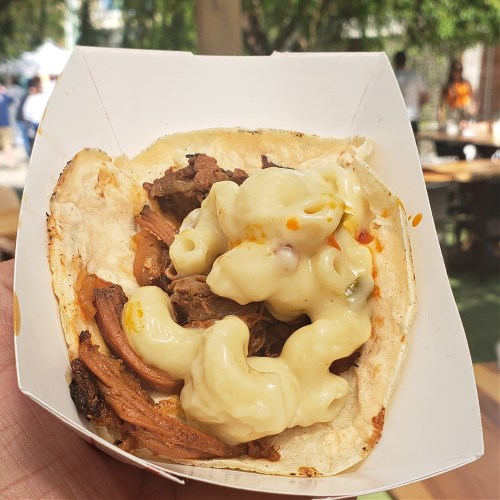A hand holding a taco filled with tender meat and creamy macaroni and cheese, set against a blurred outdoor background.