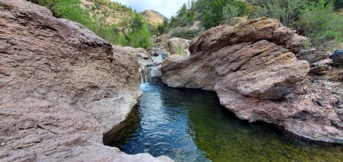 A serene rocky landscape with a small waterfall flowing into a clear pool, surrounded by greenery and hills.