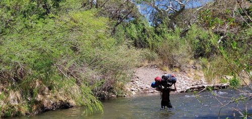 A person wades through a river, carrying a large backpack, surrounded by lush greenery and rocky banks.