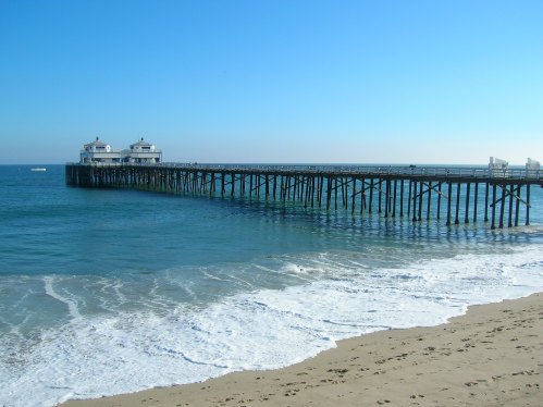 A wooden pier extends over calm blue waters, with gentle waves lapping at a sandy beach under a clear sky.