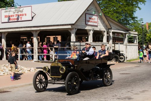 A vintage Model T car drives past a crowd outside a historic building on a sunny day.
