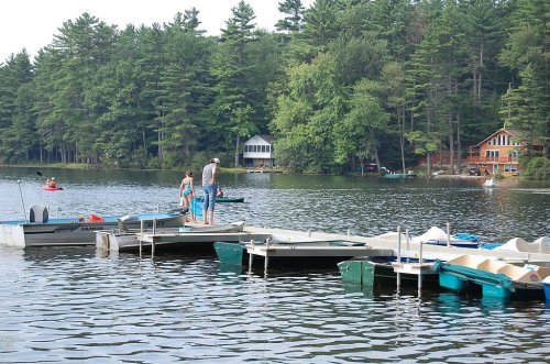 Two people stand on a dock by a calm lake, surrounded by trees and boats, with a cabin visible in the background.