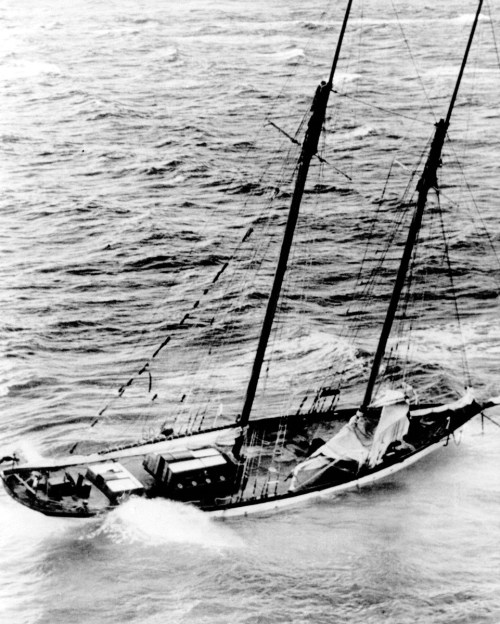 A black and white image of a sailing ship navigating rough waters, with visible sails and cargo on deck.