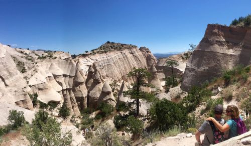 A couple sits on a rock, overlooking a stunning canyon with unique rock formations and greenery under a clear blue sky.