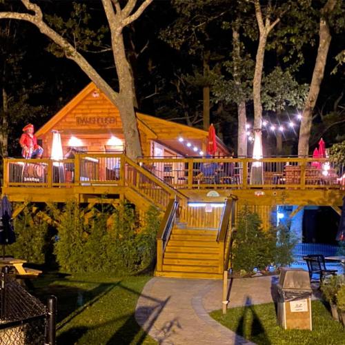 A wooden cabin at night, surrounded by trees, with outdoor seating and lights illuminating the area.