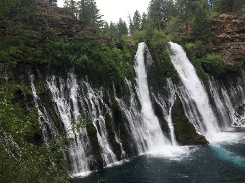A scenic waterfall cascading over rocky cliffs into a clear blue pool, surrounded by lush green trees.