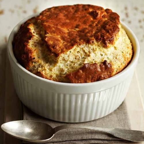 A golden-brown soufflé rises in a white ramekin, resting on a linen napkin with a spoon beside it.