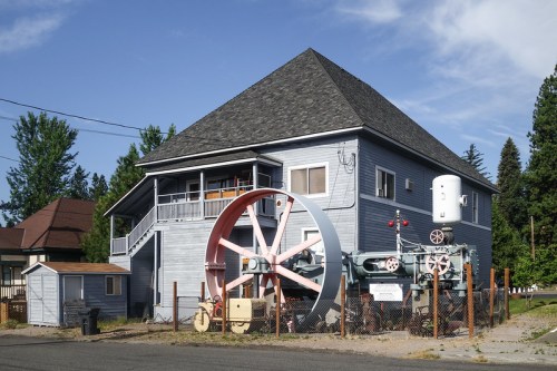 A large blue house with a distinctive wheel and machinery displayed in front, surrounded by a fence and trees.