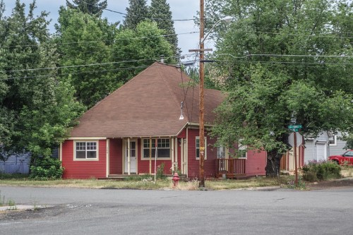 A red house with a brown roof sits at a street corner, surrounded by trees and power lines.