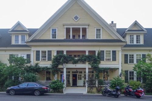 A large, yellow Victorian-style house with a porch, surrounded by greenery, featuring a car and two motorcycles parked out front.