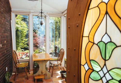 A sunlit dining area with a wooden table and chairs, surrounded by large windows showcasing a garden view.