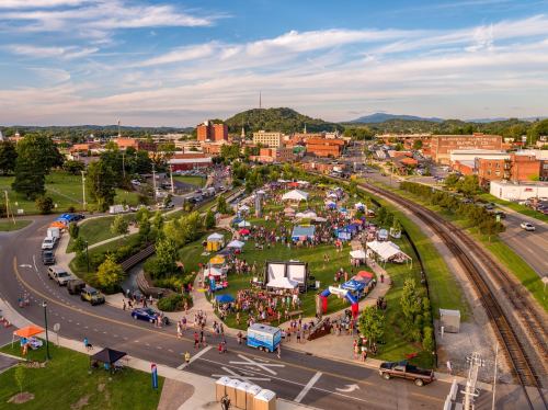 Aerial view of a vibrant outdoor festival with tents, people, and a scenic backdrop of hills and buildings.