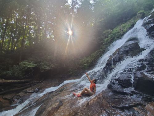 A person sits on a rocky waterfall, reaching up towards the sun shining through the trees above.