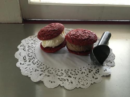 Two red velvet cookie sandwiches filled with vanilla and chocolate ice cream, placed on a lace doily with a scoop nearby.
