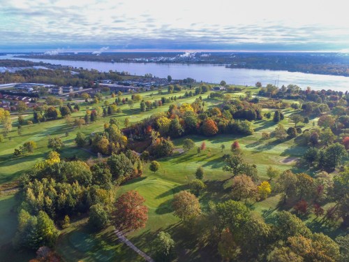 Aerial view of a lush green landscape with colorful trees, a river, and distant buildings under a cloudy sky.