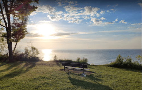 A serene lakeside view at sunset, featuring a bench on green grass and soft clouds in the sky.