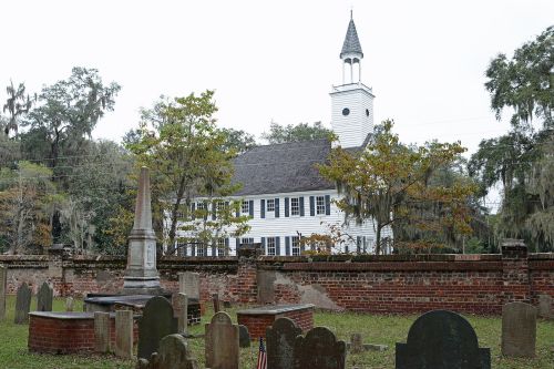 A historic church behind a cemetery with gravestones and a tall monument, surrounded by trees and moss.