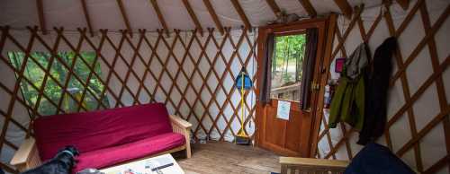 Interior of a yurt featuring a red couch, wooden furniture, and a door leading outside, with natural light coming in.