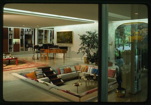 A modern living room with a sunken seating area, piano, bookshelves, and large windows showcasing greenery outside.