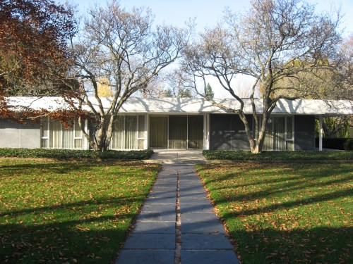 Modern house with large windows, surrounded by trees and a grassy lawn, featuring a stone pathway leading to the entrance.