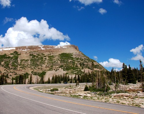 A winding road curves around a mountainous landscape with green vegetation and a blue sky dotted with clouds.