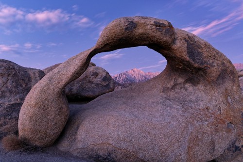 A natural rock arch framed by boulders, with mountains in the background under a colorful sky at dusk.