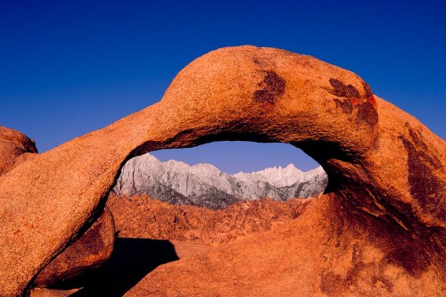 A natural rock arch frames snow-capped mountains against a clear blue sky.