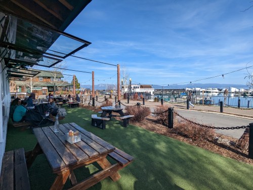 Outdoor seating area with wooden tables, overlooking a marina and blue sky, with people enjoying the view.