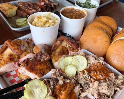 A spread of barbecue featuring grilled chicken, pulled pork, sides of macaroni, beans, and buns on a tray.