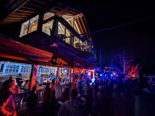 A lively outdoor scene at night with a crowd gathered near a brightly lit building and colorful lights.